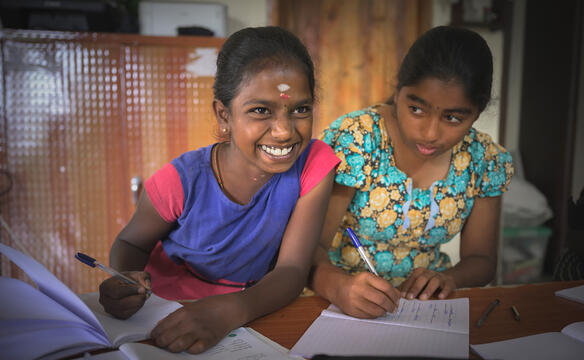 Zwei Mädchen schreiben etwas in ihre Hefte, eins lächelt in die Kamera