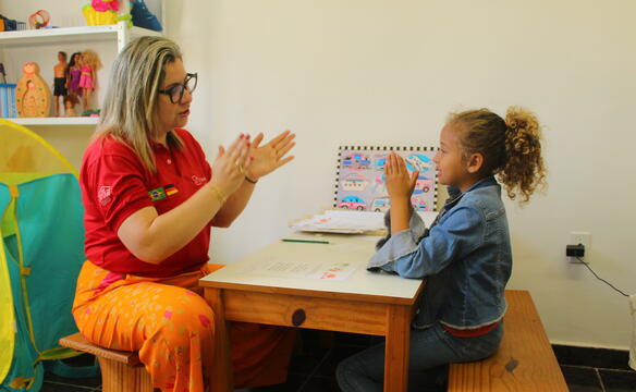 Eine Frau sitzt einem Mädchen an einem Tisch gegenüber, beide haben die Hände zum Klatschen gehoben