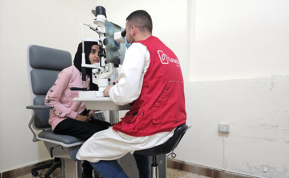 Ein Arzt mit roter humedica Weste untersucht mit einem Gerät die Augen einer Frau